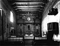 Historic interior photograph of the sanctuary at Mission San Miguel Arcángel. Beamed wooden ceilings and adobe walls protect an ornate altar, chancel, and pulpit.