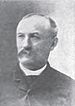 Head and shoulders of a balding white man with a mustache, wearing a dark suit coat.