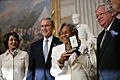 Black woman holding aloft award presented by President George W. Bush and two other dignitaries
