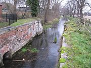 View downstream of the lock chamber build for the Sleaford Navigation, to maintain the head at the mill.  The chamber is largely brick built with stone details for load-bearing parts, and the brick is coloured with moss and lichen.  A little desultory grass covers the top sides.  There are no lower gates, the lock having been converted into a weir many years ago.  A cheap iron railing fence, painted black recently, delineates the property associated with the mill and restaurant to the left. This is a winter view and many bare trees line the banks downstream. The trunks of the nearest can be clearly seen to be covered in ivy.  The water looks clear and placid.