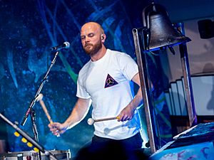 A bald, bearded man wearing a white shirt plays the timpani while a church bell is beside him