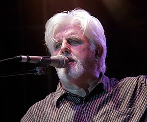 Man with silver shoulder-length hair and goatee in white v-neck top.