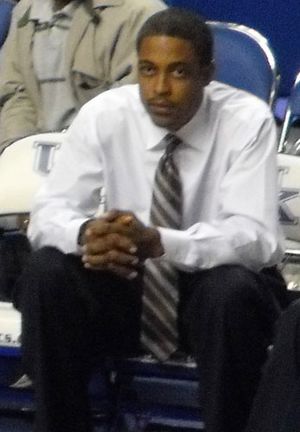 An African-American man sits with hands folded wearing a white shirt and tie