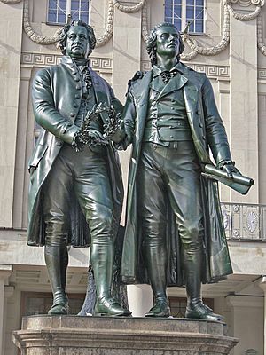 Photograph of a statue of Goethe and Schiller standing side by side, each looking forward. The statue is in front of the stone facade of an elaborate building. They are of nearly the same height. Goethe appears middle-aged; Schiller is noticeably younger. They are dressed in nineteenth century clothing. Goethe is wearing a knee-length formal coat, and Schiller is wearing a calf-length coat. Both men wear breeches. Goethe's left hand rests lightly on Schiller's shoulder; his right hand holds a laurel wreath near his waist. Schiller's right hand is nearly touching the wreath, which may suggest that Goethe is passing the wreath to Schiller. Schiller's left hand extends loosely below his waist, and grasps a rolled sheet of paper.