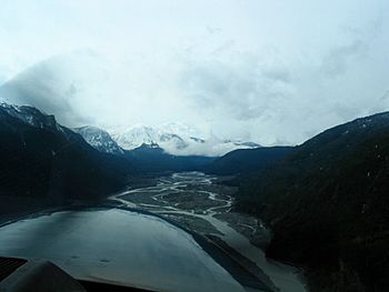 To he right and left are mountains, between which is a deep valley with numerous meandering and intersecting segments of the Iskut River proceeding into the distance. Beyond it are snow-capped mountains.