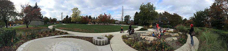 A busy park with children riding bikes, using playground equipment, and digging in a sandbox, with a blue water tower in the background.