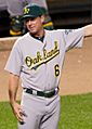 A man wearing a gray baseball uniform with "Oakland" and the number "6" written across the chest in green letters and a green cap with a yellow "A" on it stands on a baseball field
