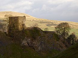 A stone tower with a wall running off to the right standing on top of a drop into a gorge. There are hills in the background.