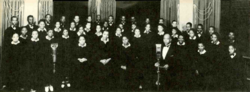 A total of 44 African-American singers, plus director Glenn Thomas Settle, posing for a publicity photo with an ornate background featuring multiple window curtains. At the front are microphones labeled "WGAR" (the originator of their radio program) and "CBS" (the network which carried the program nationally).