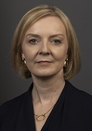 Truss facing frontward, with short blonde hair and dark clothes in front of a grey background.
