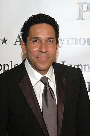 Close-up of Oscar Núñez wearing a black suit and tie with white shirt, smiling at camera
