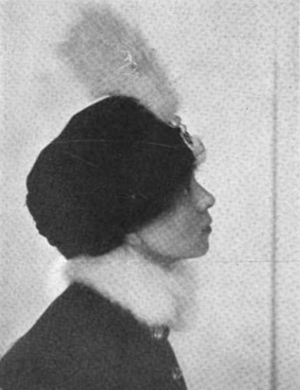 Woman in feather adorned hat and fur collared coat seen in profile