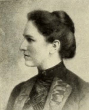 A white woman with her dark hair in a bouffant updo, wearing a dark high-collared dress and jacket with embellished lapels; portrait in profile