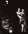 A rock band is onstage. A drumkit is on the left. A singer, Iggy Pop, sings into a microphone. He is wearing jeans and has no shirt on.