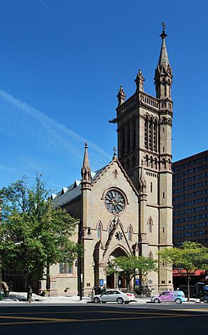 A Gothic stone church with a pointed facade and a tower on the right, with a smaller tower rising from its own right, seen from across a city street