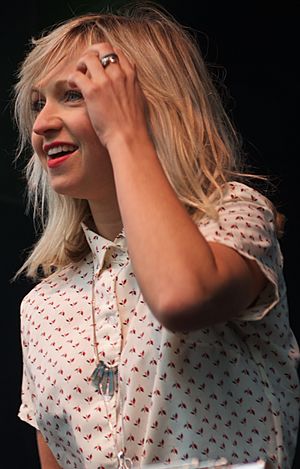 close-up of Ashleigh Ball wearing a white print blouse, holding her left hand up to her temple, with a grin on her face