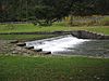 White water spills over a stone dam in a grassy setting with a highway guardrail in the background