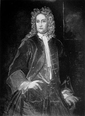 Grayscale image of a man in allonge wig, waiscoat and coat standing with hand on hip