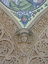 A carving of the head and wings of an angel: above the angel is the bottom of a mosaic with the label "HOPE" and a margin which has a head with flowering ivy.