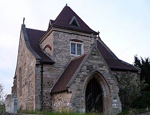A squat stone church with a gabled porch, beyond which is a low tower with a pyramidal roof, and the body of the church is behind this