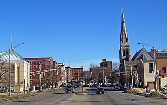 A view from the middle of an urban street looking toward a square some blocks away where a monument stands on a tall pedestal, in winter. Buildings of several stories in height line the street on either side; on the right is a church with a tall steeple.