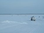 Large trucks, widely spaced, travel single-file across a frozen river.