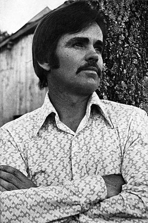 Photo portrait of a man with medium-length hair and a mustache crossing his arms and standing in front of a tree and a wooden shed