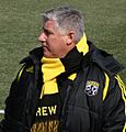 A portly man with graying hair. He is wearing a coat and scarf that are both black and yellow.