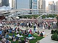 A large number of people are seated on the grass facing a bandshell with an even larger crowd across a wide sidewalk. The bandshell is framed by shiny curved metal and a trellis is over the larger crowd. Skyscrapers are in the background. 