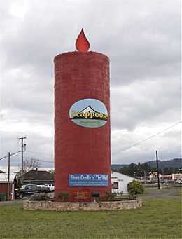 A cylindrical-shaped red tower-like structure resembling a candle sits inside a circular stone brick wall with plants and shrubs around it. A red flame-like structure sits atop the candle, and an oval-shaped logo with the word "Scappoose" in front of a drawing of a mountain rests in the center of the candle. A blue banner with the words "Peace Candle of the World" in white is draped near the bottom of the structure. In the background is a rural setting with houses and trucks below a sky with gray clouds.