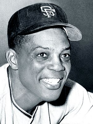 Black-and-white portrait photo of a broadly-smiling man wearing a dark-colored baseball cap with SF embroidered on it