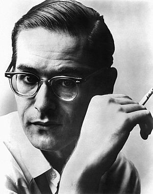 Headshot of a man with short slicked-back hair and horn-rimmed glasses smoking a cigarette