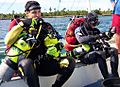 Two scuba divers in one-piece wetsuits and full scuba gear sitting in a boat, preparing to dive. Both divers are wearing tech rigs with back inflation BC and sling cylinders for decompression gas