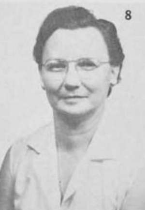 A white woman with short dark hair, wearing glasses and a light-colored blouse with a notched collar; the number 8 is in the upper right corner of the photo