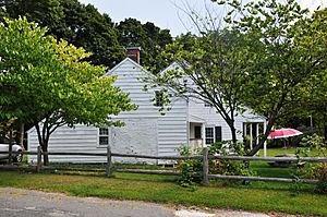 A white wooden house in two sections behind a wooden fence. Both have black pointed roofs with brick chimneys. The one on the right is slightly larger and has an open full-length porch on the right underneath the overhanging roof eave.