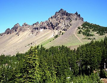 A jagged mountain with spires rises above its lightly forested slope, with a more heavily forested area in the foreground of the picture.