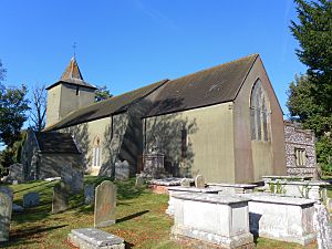 Three-quarter view of a long, low, grey church behind tombs and gravestones in a churchyard.  There is a spire-topped tower at the far end of the brown tiled roof.