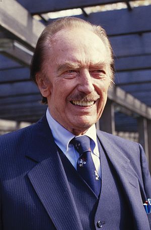 Photographic portrait of a balding blondish older man with a mustache. He is smiling, and his prominent eyebrows and lower eyelids nearly conceal his blue eyes. His right cheekbone is sunken in around the upper area of that side of the jawbone. His perfect teeth are just off-white. He is wearing a blue suit and tie.