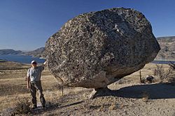 A man stands beside a balancing rock near a lake