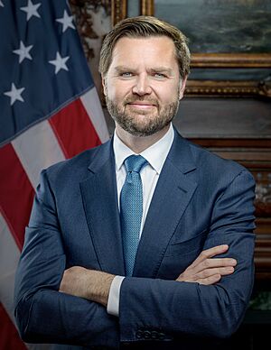 Official portrait of JD Vance, a middle-aged white man with dark hair and beard and light eyes, wearing a suit and tie, crossing his arms while standing in front of an American flag.