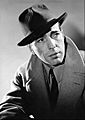 Black and white publicity photo of Humphrey Bogart—a charming white man with dark eyes and a square face, wearing a dark hat and a light-colored overcoat, around 41 years of age—in 1940.