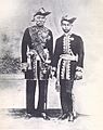 Black and white photo of older man and teenage boy standing in uniforms with bold designs and ornaments