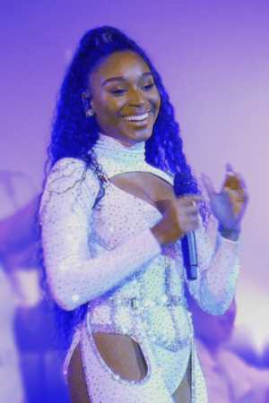 Normani holds a microphone in her hand, looking right and smiling