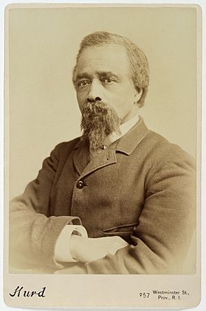 Black-and-white, bust-length portrait photograph of Edward Bannister in a cabinet card mount. He is looking directly at the camera, has his arms crossed, and is wearing a jacket and collared shirt.
