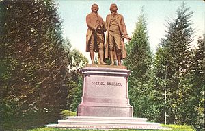 Picture postcard of a large bronze statue of two men on top of a stone pedestal. The pedestal has the engraved words "Goethe. Schiller." on its front face. The statue is surrounded by tall trees. There is printing along the bottom of the postcard that says, "1671 – Goethe Schiller Monument, Golden Gate Park, San Francisco, California" printed along the bottom.