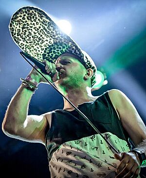 White male standing onstage, holding microphone to his mouth, wearing tank top and unusual hat with brim