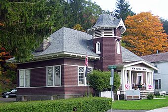 A brown wooden building with a gray peaked roof and tower and porch in front. Behind it are trees. One on the right is bright orange with autumn color