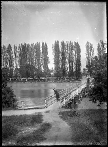 Albert Percy Godber standing on a footbridge over the Taruheru River, Gisborne, with a row of poplars on the far side. ATLIB 291677.png