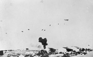 A blurry black and white photograph of an aircraft with numerous parachutes descending and a large smoke plume in the distance