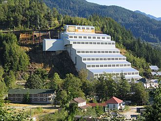 A large, white, stair-step structure built into the base of a forested mountain. It consists of eight levels, the topmost narrower than the others, each with a row of compound windows at its top subdivided into vertical groups of four. The top of the structure is connected to a pipe leading off to the left, supported by metal piers anchored into the cliff. Parallel to and above the pipe are electrical cables. The mountain rises out of the frame to the left, and behind it on the right are visible other forested mountain bases. At the foot of the building and mountain are several smaller buildings.